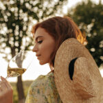 stylish young girl with foxy hairstyle in modern straw hat and cool summer outfit posing with glass